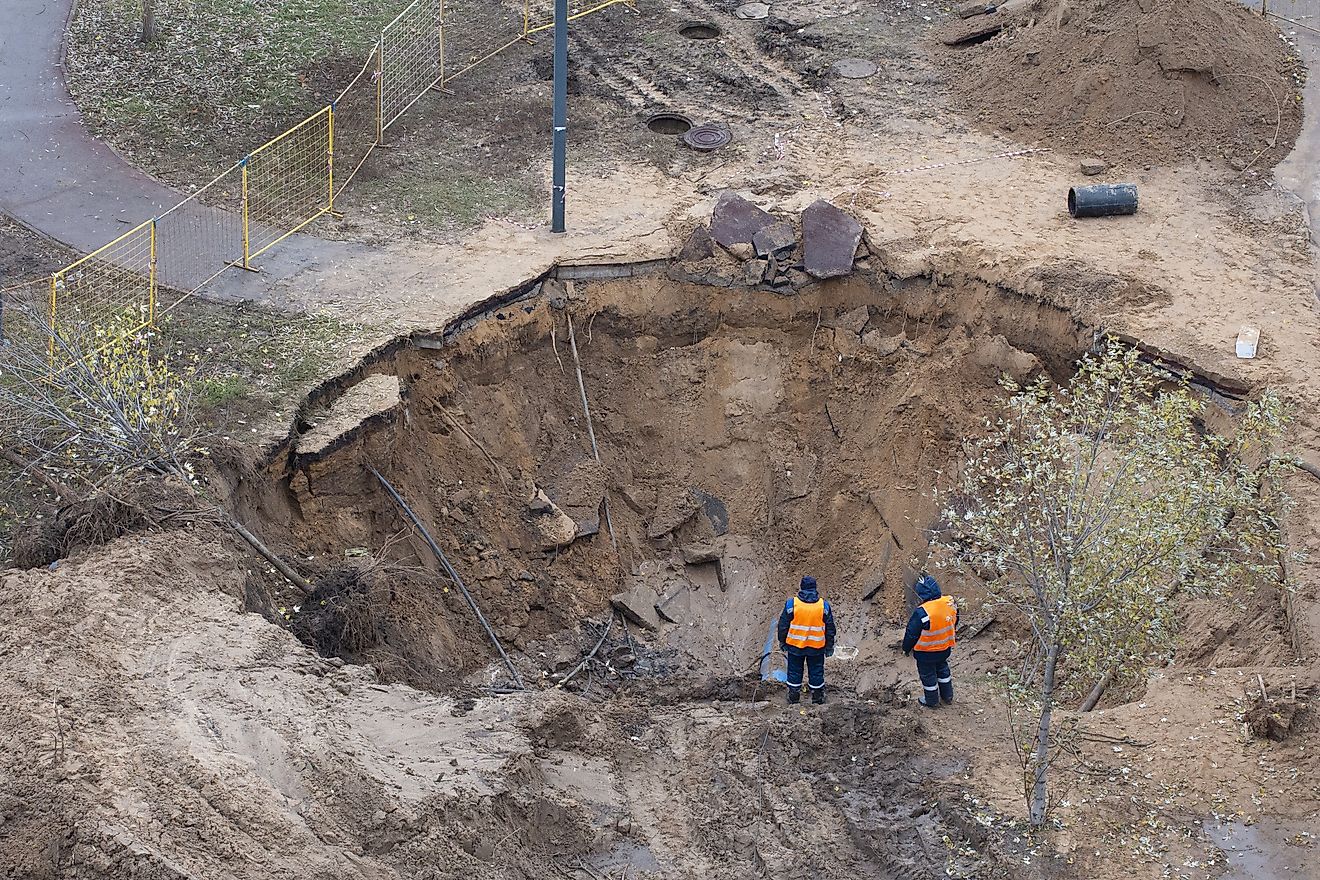 An aerial shot shows two workers in safety vests assessing a large sinkhole that has opened up near a path, disrupting the urban landscape