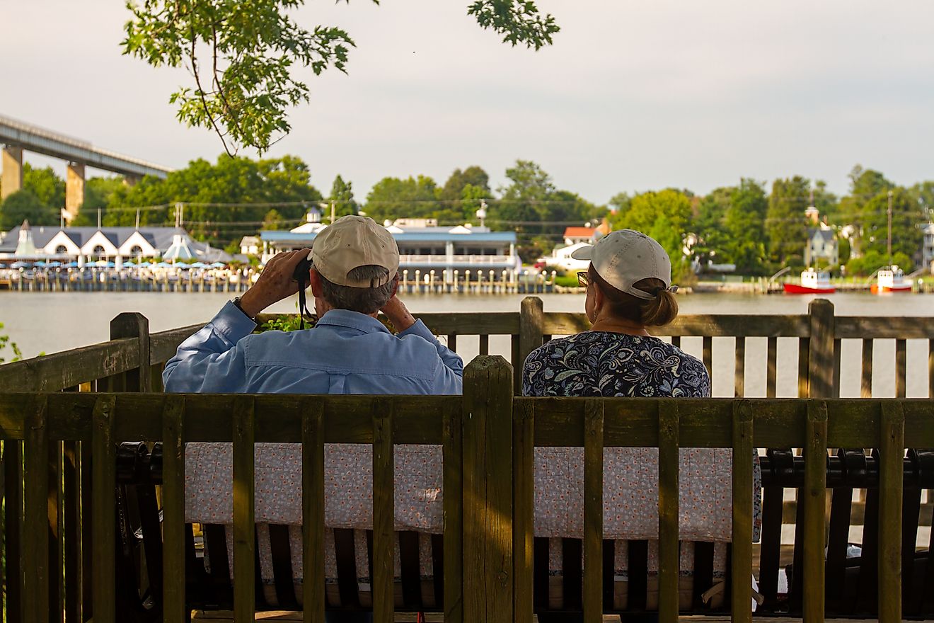 An elderly couple wearing baseball hats is sitting on a wooden outdoor bench in a garden deck by the Chesapeake and Delaware Canal. 