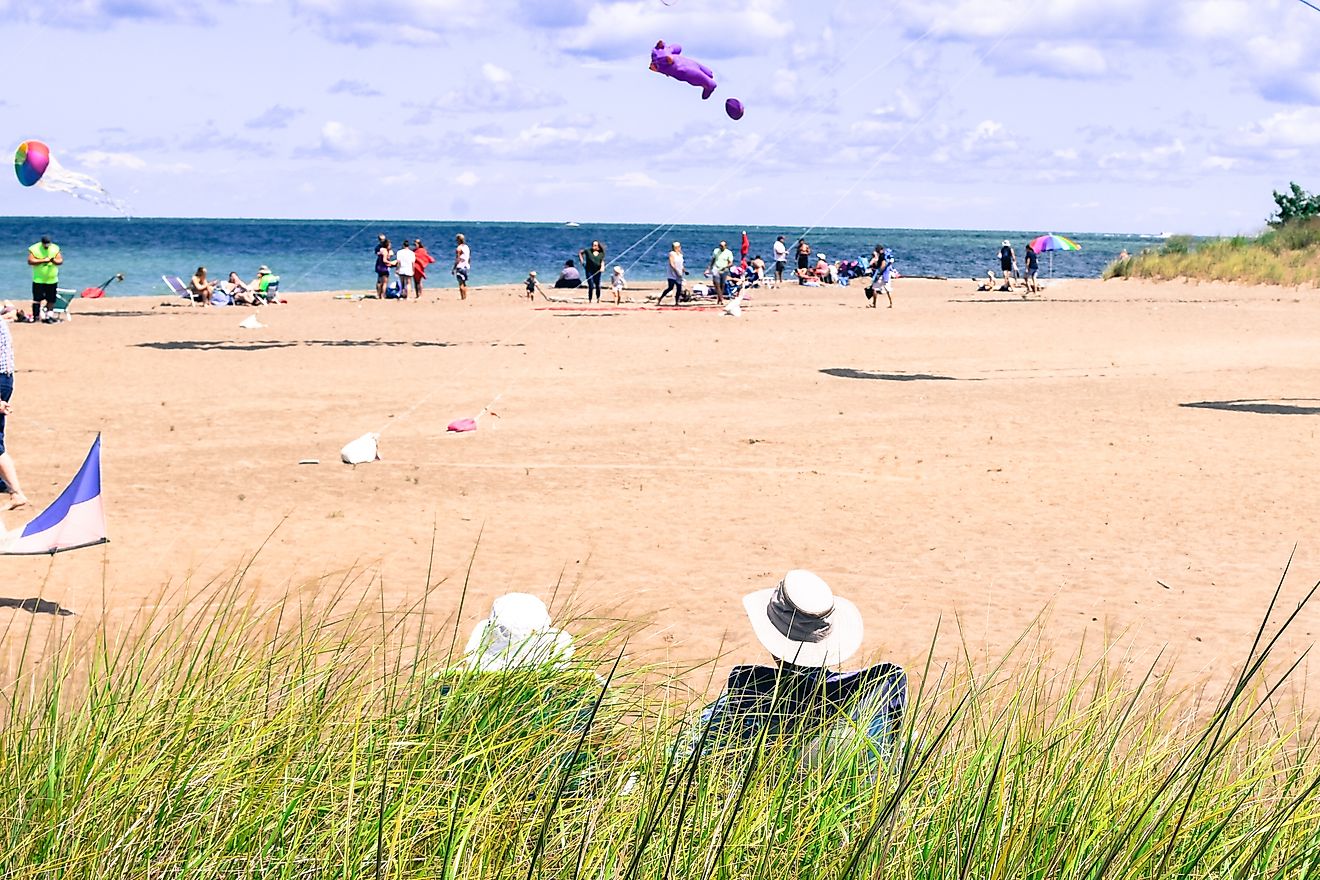 back of a couple wearing fishing sun hats sitting in lawn chairs behind tall green grass at the beach relaxing with people and kites in the background at Presque Isle State Park